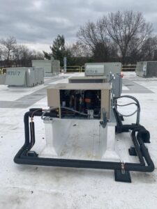 Rooftop HVAC unit with exposed coils and surrounding insulated pipes, situated on a flat roof with other RTU units visible in the background under an overcast sky.