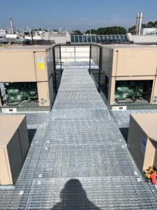 Metal walkway on a rooftop between HVAC units under clear sky, with a person's shadow visible in the foreground.