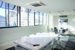 A modern conference room with a large white table, several white chairs, and floor-to-ceiling windows overlooking city buildings.