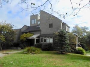 Modern two-story gray house with angular rooflines, large windows, and surrounding greenery on a grassy lawn.