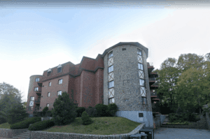 A multi-story brick and stone apartment building with rounded towers, surrounded by trees and greenery, viewed from the parking area.