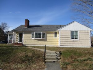 Single-story house with a gray shingle roof and mixed siding; left side painted yellow, right side partially painted white. Concrete steps lead up to the front door.