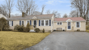 Single-story beige house with blue shutters, attached garage, front porch, and a paved driveway, set on a lawn with leafless trees in the background.