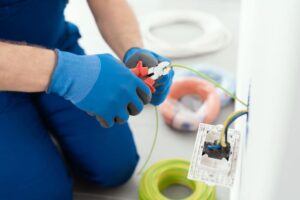Person wearing blue gloves uses pliers to strip the insulation from a green electrical wire near an open electrical outlet with various colored wires.