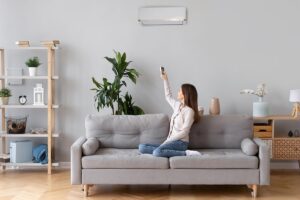 A woman sits cross-legged on a gray sofa, pointing a remote at a wall-mounted air conditioner in a modern living room with plants and shelves.