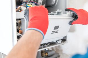 Person wearing red gloves repairing or inspecting an open gas boiler, with visible internal components and wiring.