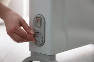 A hand adjusts the dial on a white electric heater, with power switches visible above the dial.