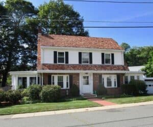 Two-story suburban house with white siding, brown shingled roof, black shutters, and a front lawn, viewed from the street.