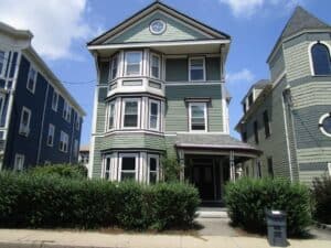 A three-story green house with white trim, bay windows, a covered front porch, and shrubs along the sidewalk under a partly cloudy sky.