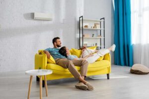A man and a woman relax together on a bright yellow sofa in a modern living room with white walls, blue curtains, and minimal decor while enjoying their wall mounted ductless air conditioner.