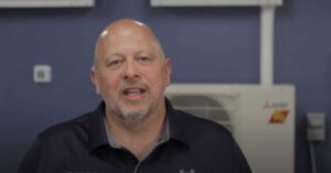 A middle-aged man with a shaved head and goatee is speaking indoors, wearing a dark collared shirt. A white wall unit and control panel are visible in the background.