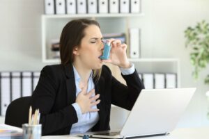A woman in business attire uses an inhaler while sitting at a desk with a laptop, holding her chest, appearing to experience breathing difficulty.