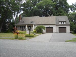 A beige two-story house with a front porch, flower garden, and attached garage, set back from the street with a paved driveway and surrounded by trees.