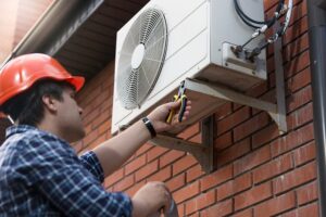 A worker in a hard hat repairs or installs an outdoor air conditioning unit mounted on a brick wall.