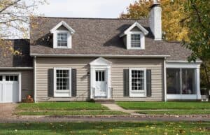 A beige, two-story house with white trim, black shutters, and a covered front entryway. The yard has green grass and trees with autumn leaves.