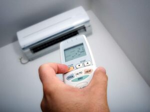A hand holds a remote control, set to 24°C, pointed at a wall-mounted air conditioning unit inside a room.