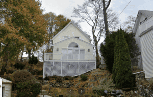 A two-story light-colored house with a large front deck and lattice skirting sits on a rocky, elevated lot surrounded by trees and shrubs.