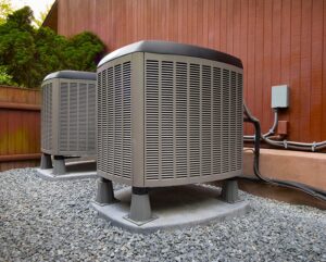 Two outdoor air conditioning units sit on concrete pads surrounded by gravel, placed next to a wooden building with pipe connections leading inside.