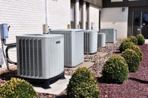 Four outdoor air conditioning units are installed beside a building, surrounded by small trimmed shrubs and decorative rocks.