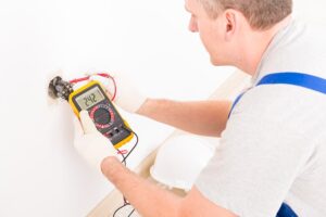 An electrician uses a digital multimeter to measure voltage in a wall socket, displaying a reading of 242 volts. A white hard hat is on the floor nearby.