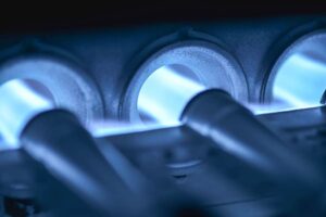 Close-up view of blue flames inside a gas furnace burner, showing multiple metal tubes with visible combustion.