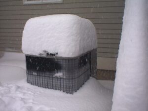 An outdoor air conditioning unit covered in a thick layer of snow beside a house during winter.
