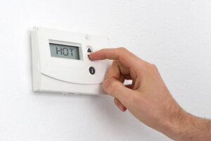 A hand pressing a button on a wall-mounted digital thermostat displaying the word "HOT" on its screen.