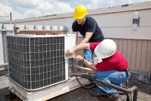 Two workers wearing hard hats perform maintenance on an outdoor air conditioning unit atop a building.