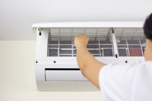 A person cleans or replaces the air filter of a wall-mounted air conditioner with the front panel open.