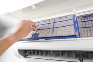 A hand is removing a dirty air filter from an open wall-mounted air conditioning unit.