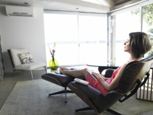 A woman in a pink dress relaxes on a lounge chair by large windows, holding a book in her lap, in a bright, modern living room.