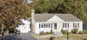 Single-story white house with a brick chimney, gray roof, and small front yard with shrubs and a red wheelbarrow on the lawn.