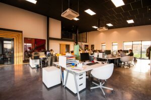 Modern open-plan office with several people working at desks, bright lighting, large windows, and contemporary furnishings.