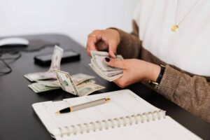 Person counting U.S. dollar bills at a desk with a notebook, pen, and smartphone nearby.