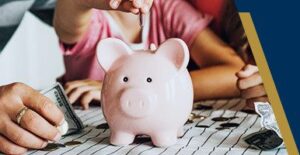 A hand drops a coin into a pink piggy bank while another hand holds cash; coins are scattered on a striped surface.