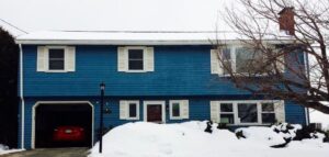 A two-story blue house with white shutters, snow-covered ground, a leafless tree, and a red car parked in an open garage.