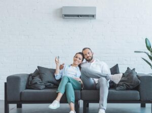 A woman and man sit together on a gray couch under a wall-mounted air conditioner; the woman holds a remote control and smiles.