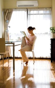 A person in pajamas sits at a dining table by a large window, reading a magazine with a mug beside them. Sunlight streams into the bright, tidy room.