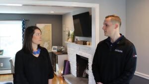 Two people stand indoors near a white fireplace, facing each other and talking in a living room with neutral colors.