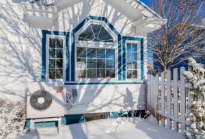A white house with blue trim, a large bay window, and an outdoor HVAC unit. Snow covers the ground, fence, and plants, with tree shadows cast on the house.