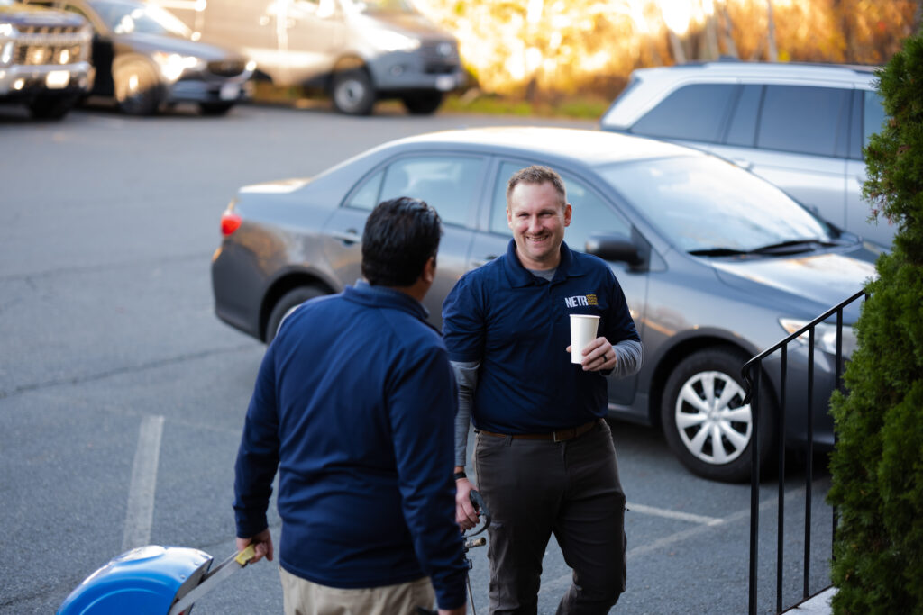 Two men in blue shirts stand and converse in a parking lot, one holding a coffee cup and the other a hard hat, likely discussing a Plumbing Repair New England project. Cars are parked in the background.
