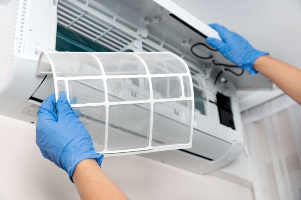 Person wearing blue gloves removing an air filter from a wall-mounted Mitsubishi ductless heat pump for cleaning or maintenance.