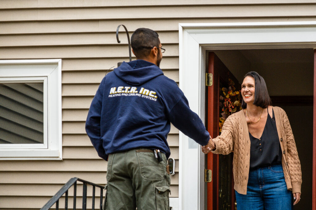 A person wearing a "N.E.T.R. Inc Heating and Cooling Systems" hoodie shakes hands with a woman at a house entrance.