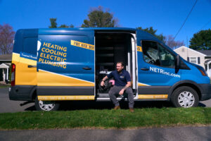 A man in work attire sits on the step of a parked NETR Inc. service van labeled for heating, cooling, electric, and plumbing services on a residential street.