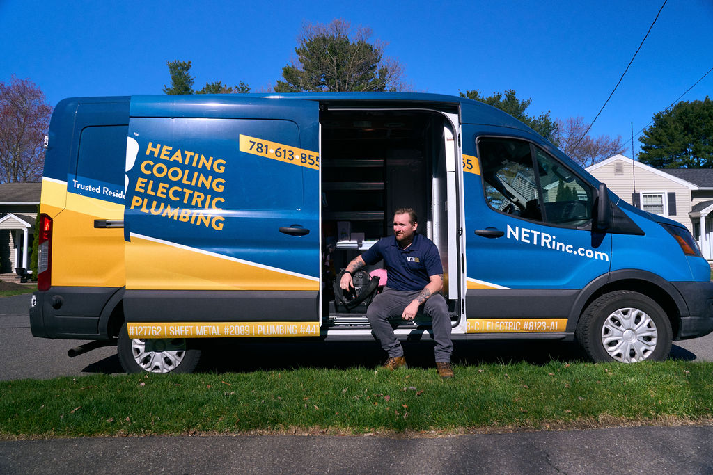 A man in work attire sits on the step of a parked NETR Inc. service van labeled for heating, cooling, electric, and plumbing services on a residential street.