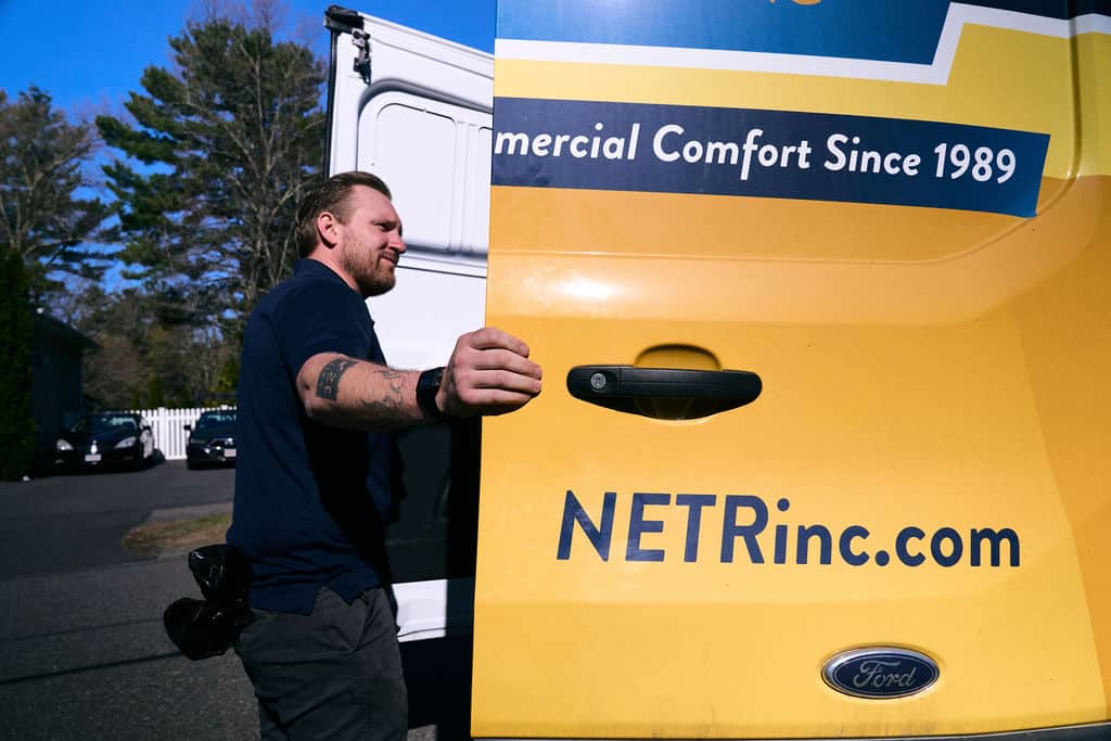 Man with tattoos closes the door of a yellow NETRinc.com service van, with the slogan "Commercial Comfort Since 1989" visible, outdoors on a sunny day.